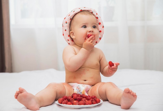 süßes beflecktes baby in einem hut mit bemalten erdbeeren, die beeren essen