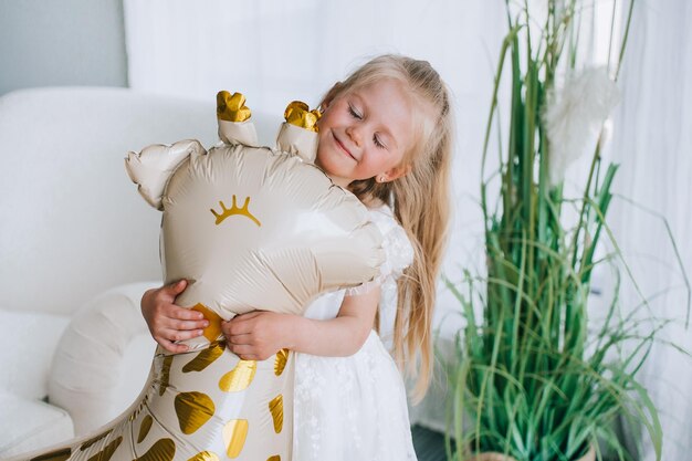 Süßes blondes kaukasisches kleines Mädchen im weißen Kleid, das zu Hause einen Spielzeuggiraffenballon mit geschlossenen Augen umarmt