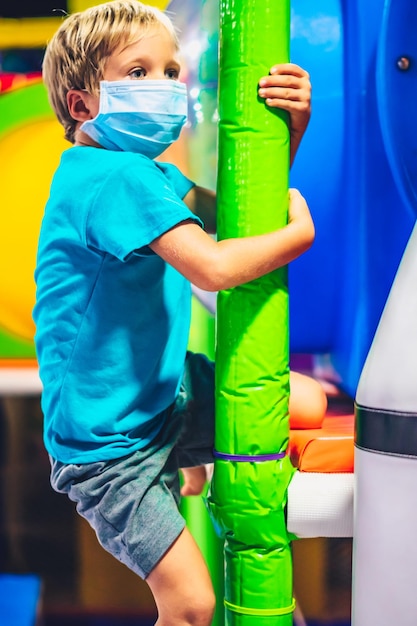 Süßes blondes Kind Junge Vergnügungsspielplatz Unterhaltung Indoor Center wickelt Arme um weiche Säule halten Hände klettern Blick weit weg Interesse warten tragen blaue Schutzmaske Anti COVID Coronavirus