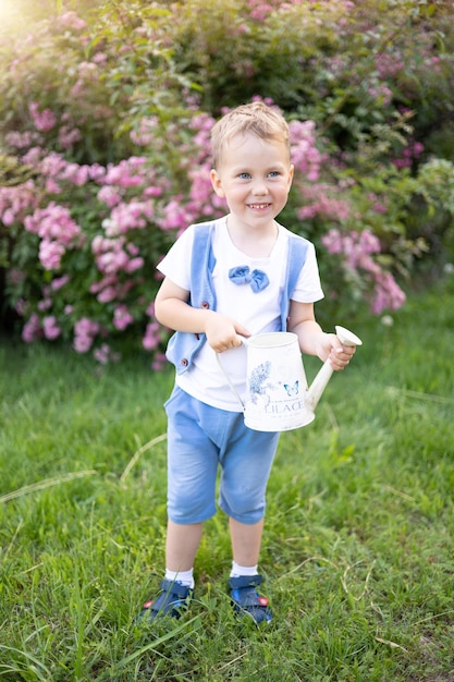 Süßes braunhaariges Baby mit blauen Augen im Sommer ein heller sonniger Tag das Kind lacht hält eine Gießkanne gießt Blumen und kümmert sich um die Natur