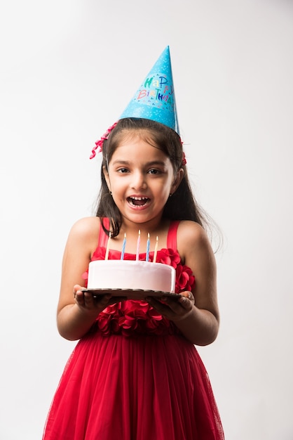 Süßes entzückendes kleines indisches oder asiatisches kleines Mädchen, das Geburtstag feiert, während es Erdbeerkuchen hält und Kerzen am Tisch bläst oder isoliert auf weißem oder rotem Hintergrund steht