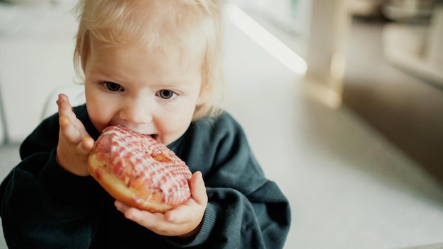 Süßes Essen und ein schönes kleines Mädchen mit blonden blauen Augen isst köstliche frische Donuts