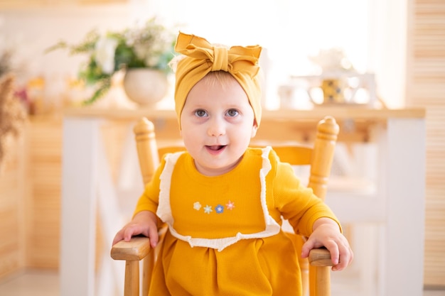 Süßes, gesundes Mädchen in gelbem Baumwollkleid, das auf einem Hochstuhl in der Küche des Hauses sitzt und Kinder mit Babynahrung füttert