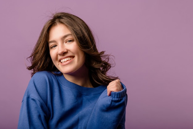 Süßes junges kaukasisches Mädchen mit welligem dunklem Haar in blauem Pullover lächelt vor violettem Hintergrund Freizeit-Lifestyle- und Beauty-Konzept