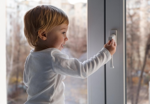 Süßes kaukasisches blondes Baby, das auf der Fensterbank lächelt, versuchen Sie Opentouch-GriffschlossKleinkindsicherheit