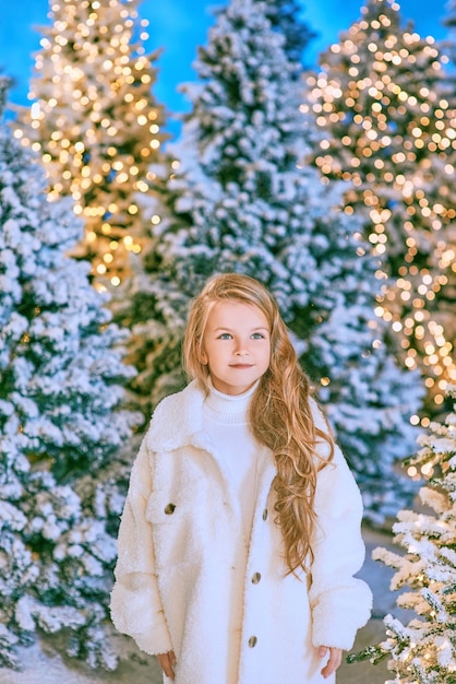 süßes kaukasisches blondes mädchen im weißen öko-pelzmantel, der im winterweihnachtswald mit lichtern spazieren geht.