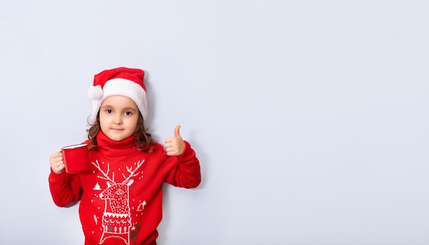 Süßes kaukasisches Kindermädchen in roter Weihnachtsmütze mit rotem Becher oder Tasse leerer Kopierraum für Design oder Text Banner Weihnachtsmockup-Vorlage