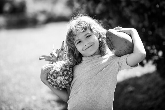 Süßes Kind mit Ananas und Kokosnuss im tropischen Garten Frische tropische Früchte für Kinder Gesunder Lebensstil mit frischen Bio-Tropenfrüchten