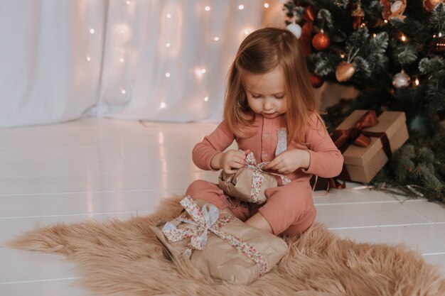 süßes kleines blondes Mädchen im rosa Pyjama öffnet Weihnachtsgeschenke unter dem Weihnachtsbaum