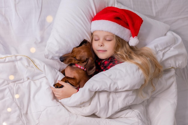 süßes kleines blondes Mädchen in Weihnachtsmütze und Schlafanzug schläft mit ihrem geliebten Hund im Bett