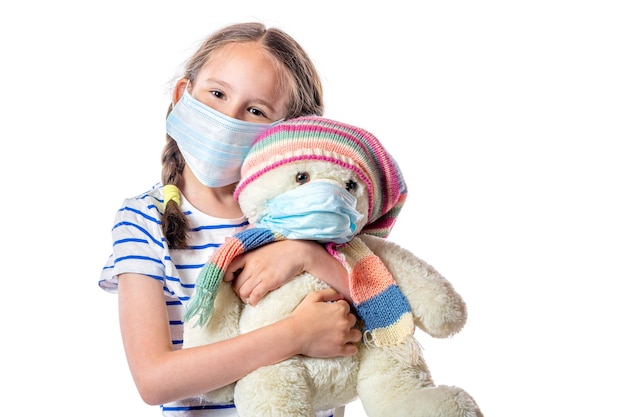 Süßes kleines europäisches Mädchen, das eine hygienische Einweg-Gesichtsmaske trägt, die einen Teddybären mit Maske hält, die auf weißem Hintergrund isoliert ist. Verhinderung der Verbreitung des Coronavirus COVID-19-Konzepts.