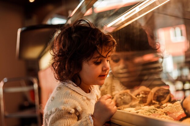 Süßes kleines lockiges Mädchen 3 Jahre alt mit einer jungen rothaarigen Mutter in der Bäckerei an der Theke wählt Gebäck Cafe