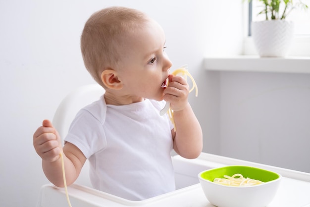 süßes kleines Mädchen, das Spaghetti-Nudeln isst, die im Kinderstuhl auf der weißen Küche sitzen