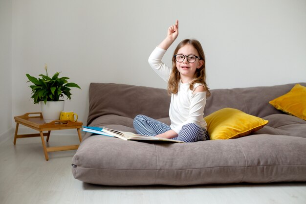 süßes kleines mädchen in brillenjeans und einem weißen rollkragen bereitet sich auf die prüfungen vor, die auf dem bett sitzen