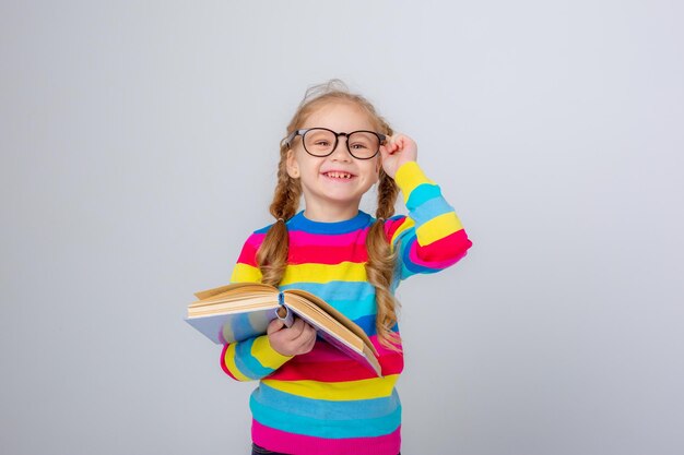 süßes kleines mädchen in einem bunten pullover und einer brille hält ein buch auf weißem hintergrund