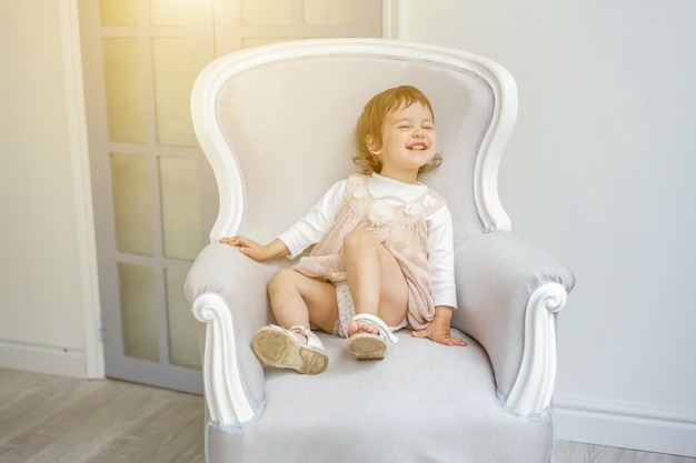 Süßes kleines Mädchen in pastellrosa Kleid zu Hause, das auf einem modernen gemütlichen grauen Stuhl sitzt und sich im weißen Wohnzimmer entspannt. Kindheit, Vorschule, Jugend, Entspannungskonzept