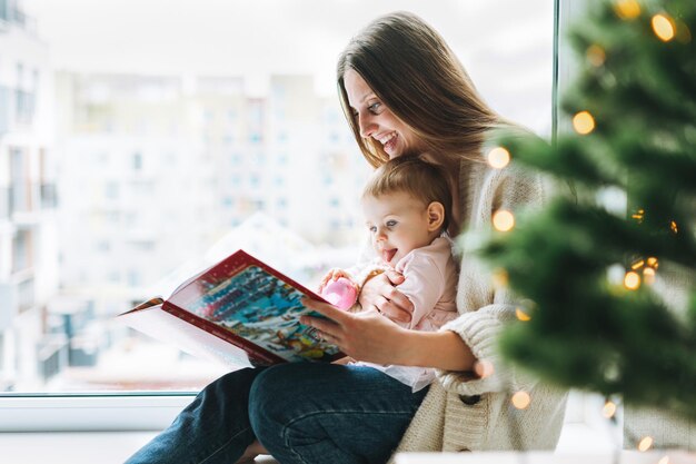 Süßes kleines mädchen in rosa kleid mit ihrer mutter junge frau liest buch im zimmer zu hause weihnachtszeit im haus frohes neues jahr