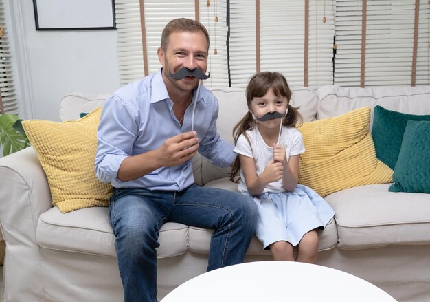 Süßes kleines Mädchen und ihr hübscher Vater mit künstlichem Schnurrbart im Wohnzimmer schauen in die Kamera