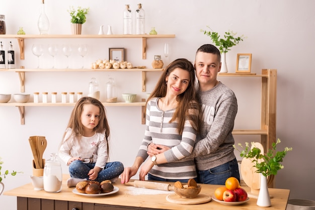 süßes kleines Mädchen und ihre schönen Eltern bereiten Essen zu und lächeln, während sie in der Küche zu Hause kochen. Den Teig auf Pfannkuchen und Brötchen kneten