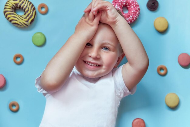 Süßes lächelndes Kind auf Donuts und französischen Macarons auf blauem Hintergrund Konzept des National Donut Day Konzept der glücklichen Kindheit Ansicht von oben