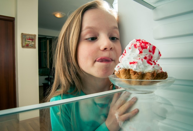 Foto süßes mädchen, das die zunge raussteckt, während es auf die eiscreme-torte auf dem teller im kühlschrank schaut