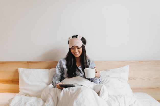 Süßes Mädchen im blauen Schlafanzug mit Lächeln liest mit einer Tasse Tee im Bett liegend