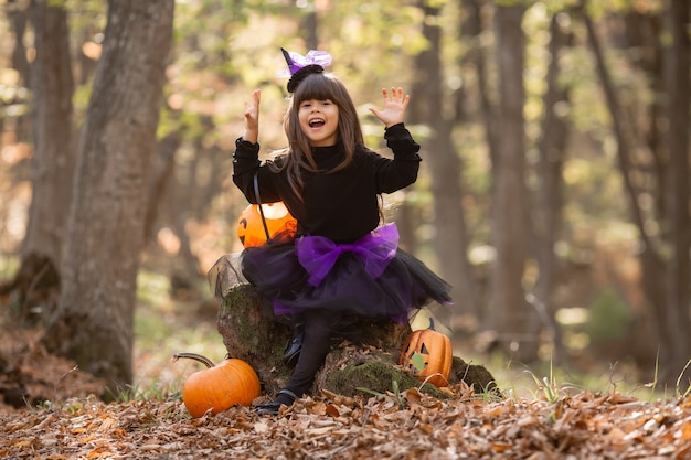 süßes Mädchen im Hexenkostüm sitzt auf Baumstumpf im Herbstwald mit Kürbislaterne Jack halloween