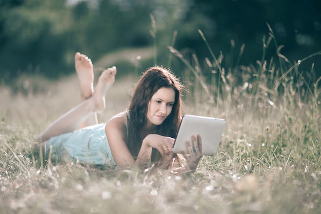 süßes Mädchen mit digitaler Tablette, die am Sommertag auf dem Rasen liegt. Mensch und Technik