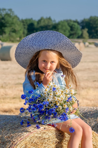 Süßes Mädchen mit Hut und einem Strauß blauer Blumen sitzt auf einem Heuhaufen