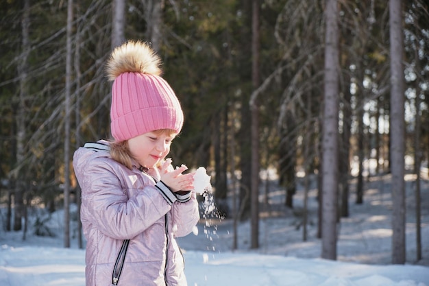 Süßes Mädchen mit Hut und Jacke hat Spaß mit Schneeflocken im Winter bei sonnigem Wetter in ihren Handflächen