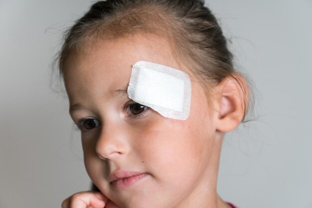 Süßes, mutiges, liebenswertes, dunkelhaariges kleines Mädchen mit Patch-Bandage im Gesicht, das der Kamera gezeigt wird Kinderschutz