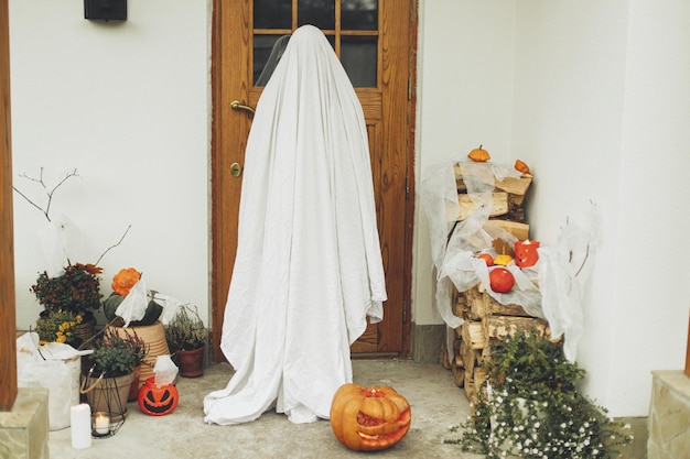 Süßes oder Saures. Gruseliges Gespenst steht vor dem Haus mit Jack-O-Laterne, Kürbis, Spinnennetz, Spinnen, Fledermäusen und Kerze. Person, die als Geist an der Halloween-Frontdekoration verkleidet ist. Fröhliches Halloween