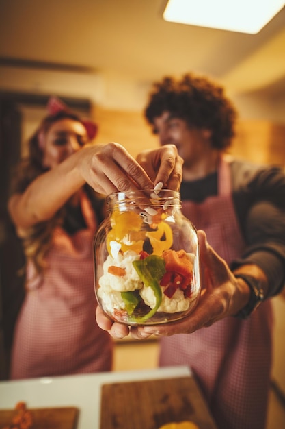 Süßes Paar legt Gemüse in ein Glas und lächelt, während es in der Küche zu Hause Gurken macht.