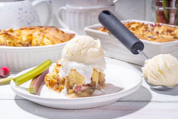 Süßes Rhubarb-Beeren-Cobbler-Torte, hausgemachter Schwammkuchen mit zuckerhaltigem Rhubarb und Erdbeeren, serviert mit Vanille-Eis-Kugel auf einem sonnigen weißen Holztisch in der Küche