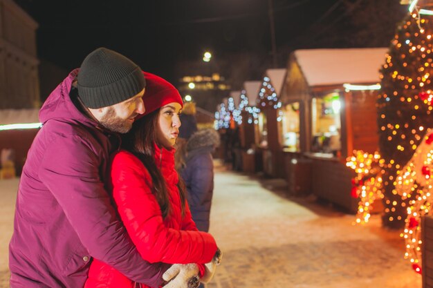 Süßes romantisches Liebespaar steht in enger Umarmung während der Neujahrsfeier in der Innenstadt, in der Nähe des Weihnachtsmarktes. Große Menge an Lichtern und Beleuchtung auf Holzhäusern im Hintergrund