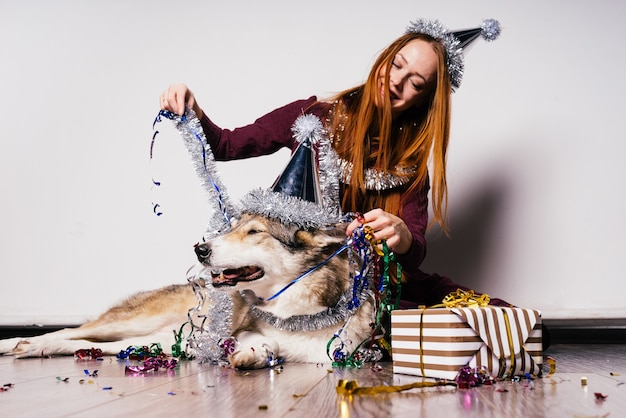 Süßes rothaariges Mädchen mit einer Mütze auf dem Kopf sitzt mit ihrem Hund auf dem Boden, feiert das neue Jahr und Weihnachten, silbernes Lametta und Geschenke