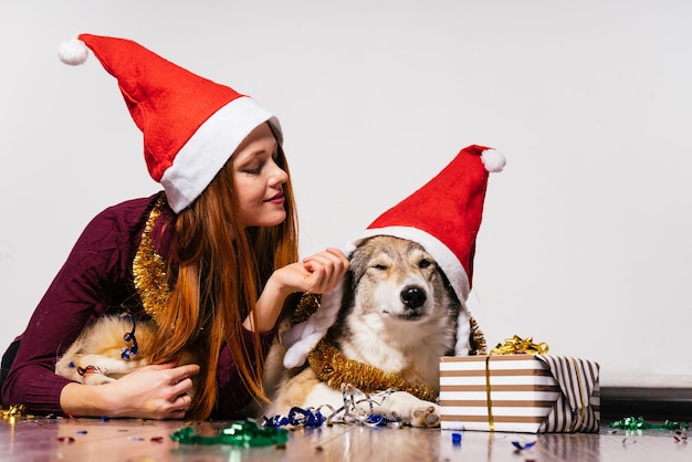 Süßes rothaariges Mädchen mit einer roten Neujahrsmütze auf dem Kopf, das mit ihrem Hund auf dem Boden liegt und auf Weihnachten wartet