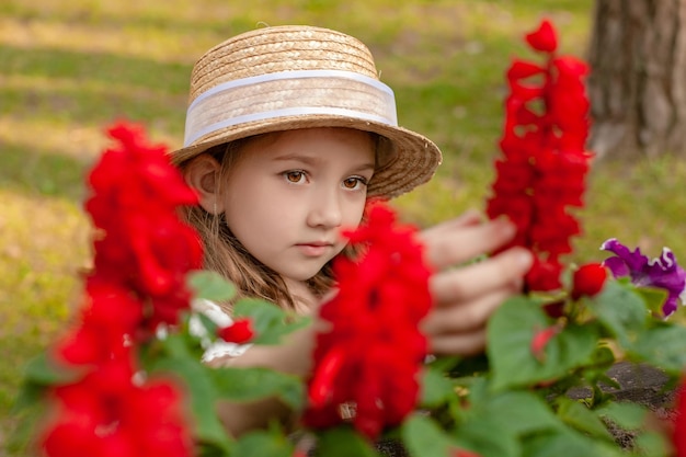 Süßes, verträumtes, braunäugiges jugendliches Mädchen mit Weidenstrohhut, das blühende rote Blumen bewundert, während es im Sommerpark-Nahaufnahmeporträt mit selektivem Fokus spazieren geht