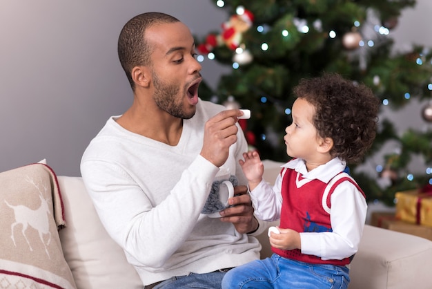 Süßigkeiten essen. Sich um einen gut aussehenden positiven Vater kümmern, der einen Marshmallow hält und ihn seinem Sohn anbietet, während er Zeit mit ihm verbringt