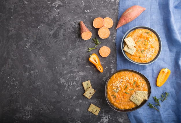 Süßkartoffel- oder Batata-Cremesuppe mit Sesam und Snacks in blauen Keramikschalen auf einer schwarzen Betonoberfläche. Draufsicht, Kopierraum.