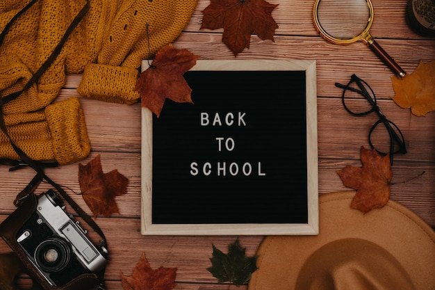 Suéter de chapéu flatlay de acessórios de outono deixa placa de carta de madeira de bordo com texto De volta à escola óculos câmera retro