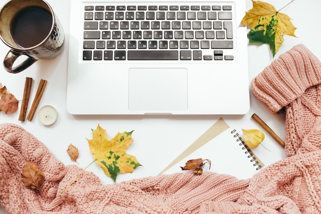 Suéter de malha, xícara de café, laptop, caderno, folhas de outono, paus de canela, velas em fundo branco. Composição de outono. Espaço de trabalho da mesa do escritório doméstico. Camada plana, vista superior, espaço de cópia