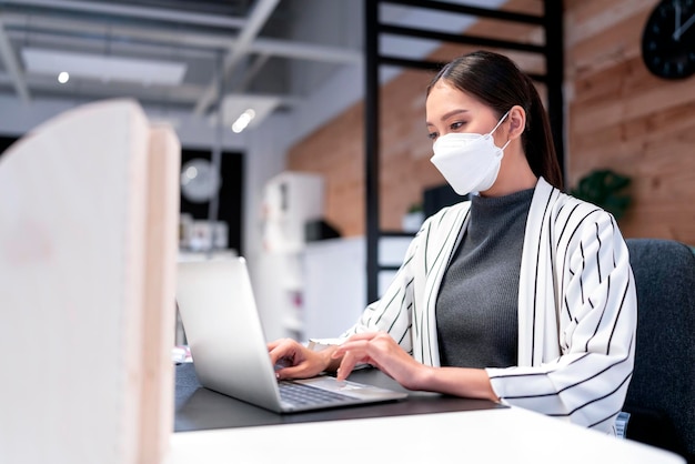 Suéter feminino asiático atraente trabalhando enquanto usava máscara de proteção contra vírus trabalhando em espaço de co woking que tem partição de plástico distanciamento social para assento novo estilo de vida normal