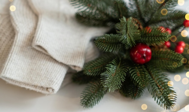 Suéter de lana blanquecino de punto doblado pequeñas piñas ramita de enebro verde guirnalda de luces doradas en una mesa de madera junto a la ventana Acogedora noche de invierno Año Nuevo Ambiente mágico de Navidad Estilo escandinavo