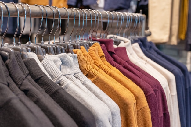 Suéteres y jerseys en diferentes colores, negro, gris, blanco y carmesí cuelgan de una percha en una tienda de ropa en una fila. colección estacional otoño e invierno
