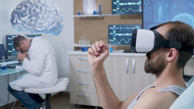 Sujeto del experimento acostado en una cama de laboratorio usando auriculares de realidad virtual, viendo una visualización mental AR holográfica mientras el científico monitorea la actividad cerebral, contribuyendo al desarrollo del campo de la neurología