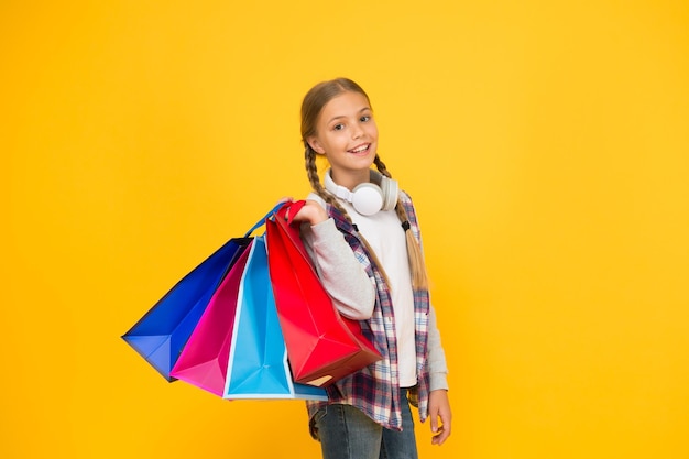 Sumérjase en las compras. Niño feliz con bolsas de papel. Sonrisa de niña con bolsas de la compra sobre fondo amarillo. Preparación y celebración navideña. Compras y rebajas el viernes negro. Consumidor adicto.