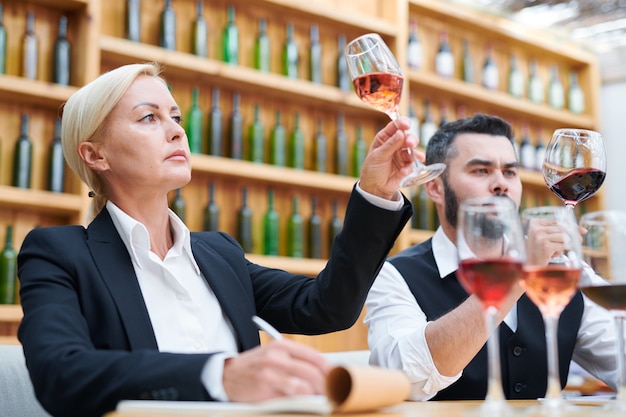 Sumiller femenino y su colega en ropa formal mirando vino en bokals mientras examina su color en el trabajo