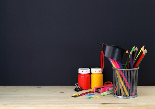 Suministros de papelería coloridos en mesa de madera y sombrero graduado contra tablero negro