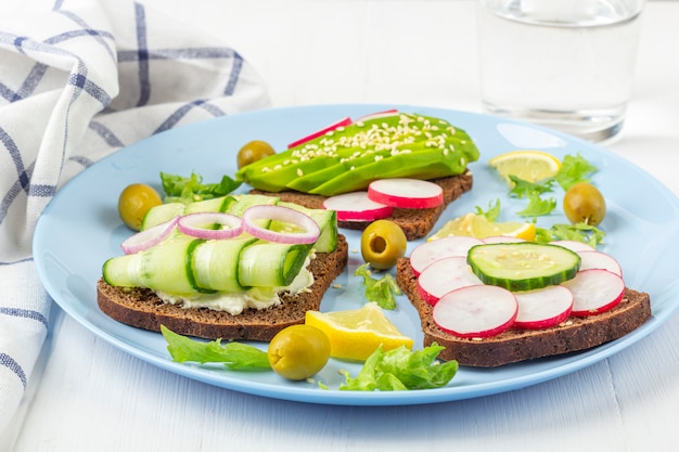 Súper alimento abierto sandwich vegetariano con diferentes ingredientes: aguacate, pepino, rábano en plato y vaso de agua sobre fondo blanco. Alimentación saludable. Texto de comida orgánica y vegetariana